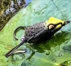A close-up of a frog-style fishing lure resting on green water lily leaves in a pond. The lure has a bright yellow hood, a black body with a raised scale pattern, and a dark tail.